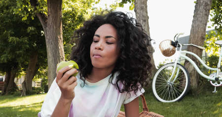 young african american woman eating fresh apple near bike in park