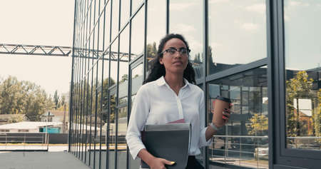 curly african american businesswoman with folders and paper cup near building