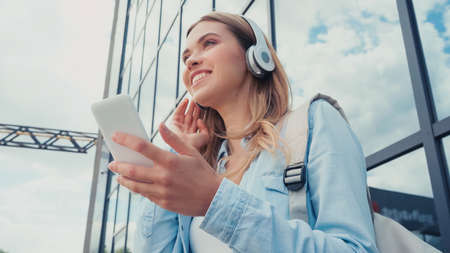 low angle view of happy woman using smartphone and listening music in wireless headphones near buildingの写真素材
