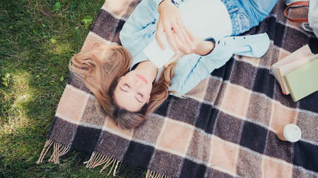 Top view of happy student using smartphone near coffee to go and books on blanket