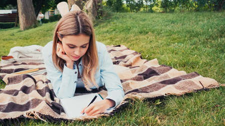 student writing in textbook while lying on blanket in park