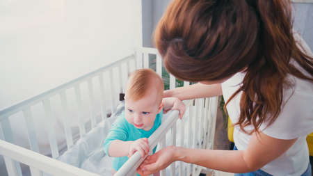 brunette woman standing near infant son in baby cribの写真素材
