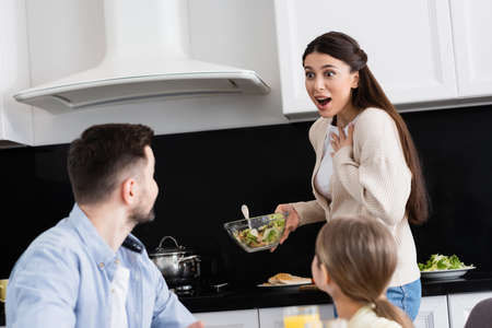amazed woman touching chest while holding vegetable salad near blurred familyの写真素材
