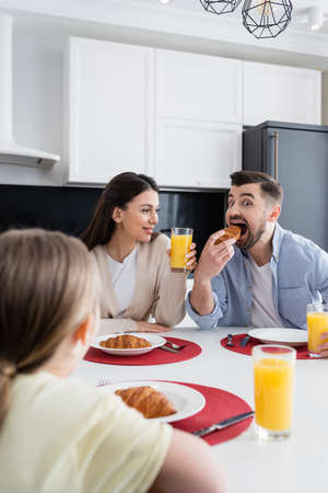 cheerful man biting growing near wife and blurred daughter during breakfastの写真素材