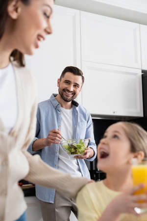 smiling man mixing vegetable salad near blurred wife and daughter in kitchen