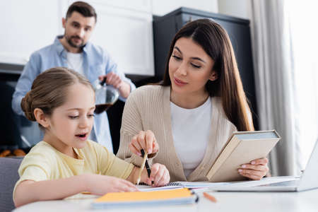 woman pointing with pencil while helping daughter doing homework near blurred husbandの写真素材