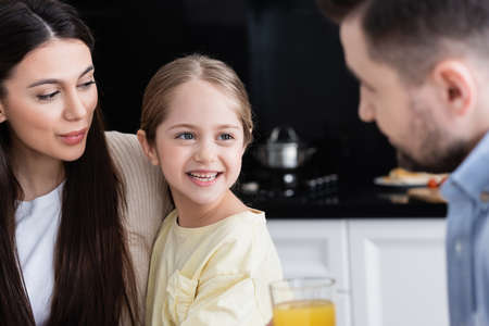 joyful girl smiling near mother and blurred father with orange juice