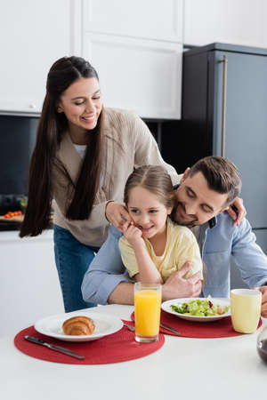 man embracing daughter during breakfast near happy wife