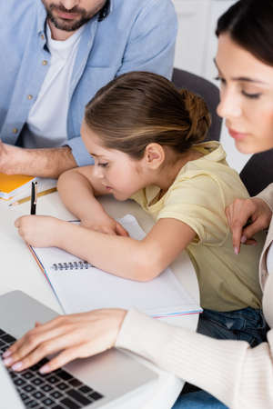 girl writing in notebook near dad and blurred mother using laptopの写真素材