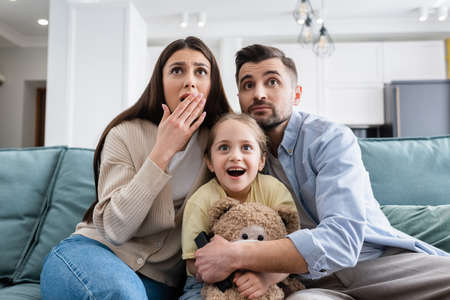 amazed girl watching movie near shocked parents and teddy bearの写真素材