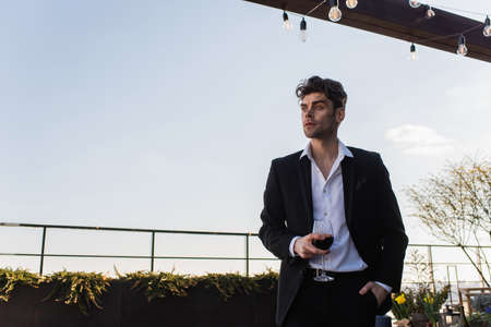 elegant man in suit holding glass of red wine and standing with hand in pocket on terraceの写真素材