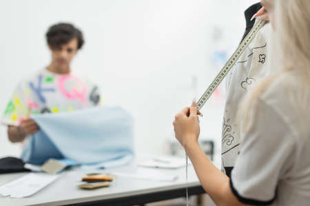 fashion designer measuring clothes on mannequin near blurred colleague holding tissue sampleの写真素材