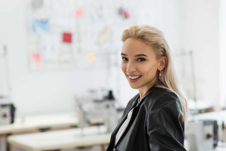 joyful, trendy fashion designer smiling at camera in tailor shop