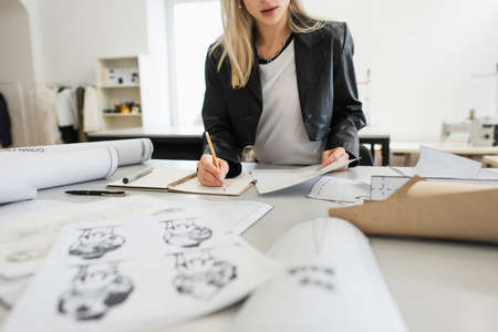 cropped view of fashion designer writing in notebook near sketches on blurred foregroundの写真素材