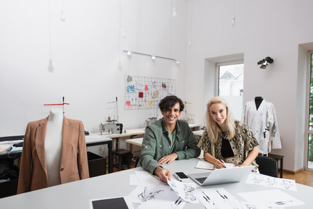 smiling fashion designers looking at camera near devices and sketches at workplaceの写真素材