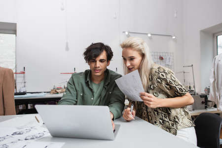 young designer looking at laptop near colleague and drawings in fashion atelier