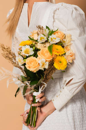 cropped view of young woman in dress holding bouquet of flowers behind back on beigeの写真素材
