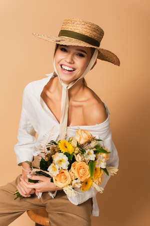 joyful young woman in straw hat holding bouquet of different flowers on beigeの写真素材