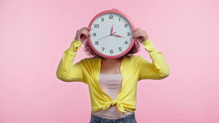 young woman covering face with clock isolated on pink