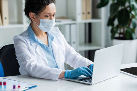 African american scientist in medial mask using laptop near digital tablet and blurred test tubes