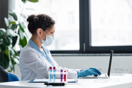 Side view of african american scientist in medical mask and latex gloves using laptop near blurred...