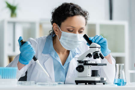 African american scientist in medical mask holding electronic pipette and looking through microscope in laboratoryの写真素材
