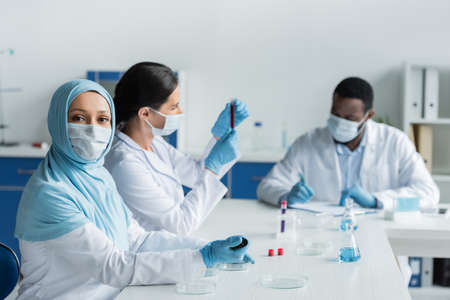 Muslim scientist in medical mask holding pipette near petri dishes and blurred colleagues in labの写真素材