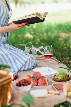 Cropped view of woman reading book near food and wine in parkの写真素材