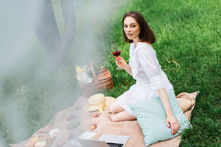 Woman with wine sitting on blanket near laptop and food during picnic in park
