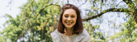 Smiling woman looking at camera in park during summer, banner