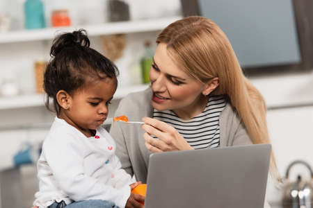 happy mother feeding adopted african american daughter with jello near laptop
