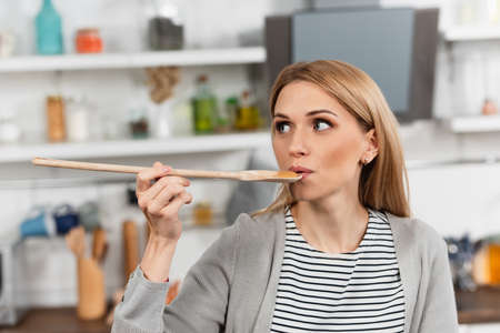 woman holding wooden spoon and trying food while cooking in kitchenの写真素材