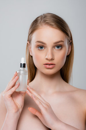 pretty woman with bare shoulders holding bottle with serum isolated on gray