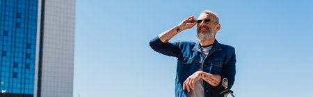 cheerful man adjusting sunglasses near e-scooter in urban street, banner