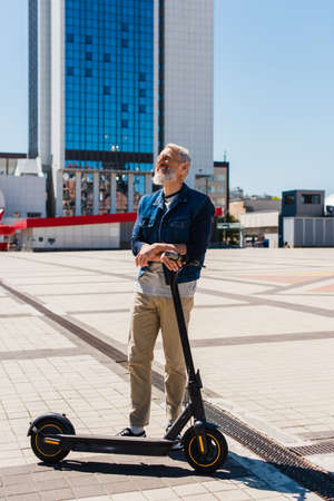 full length of cheerful man standing near e-scooter in urban city