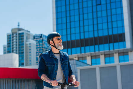 positive and mature man in helmet and sunglasses riding electric scooter on street in cityの写真素材
