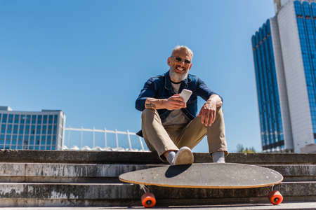 pleased middle aged man in sunglasses sitting on stairs near longboard while using cellphone on urban streetの写真素材