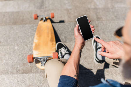 top view of tattooed mature man using smartphone with blank screen near longboard on stairs