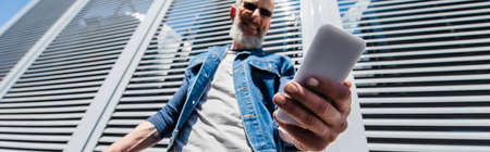 low angle view of pleased middle aged man in sunglasses using smartphone outside, banner