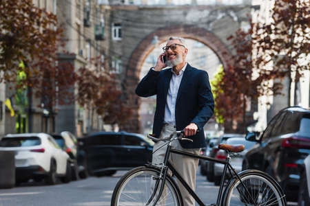 joyful middle aged man in blazer and glasses talking on smartphone and standing near bicycle on modern urban streetの写真素材