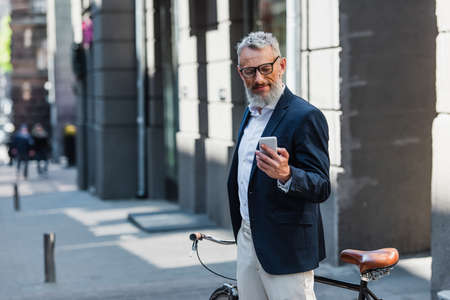 middle aged man in blazer and glasses looking at smartphone and standing near bicycle on streetの写真素材