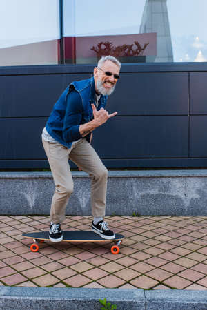 full length of happy man with open mouth riding longboard and showing rock sign on urban streetの写真素材