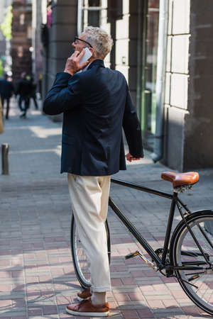 middle aged man in blazer and glasses talking on smartphone and standing near bicycle on modern urban streetの写真素材