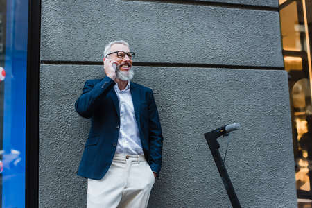 happy mature businessman in blazer talking on cellphone near e-scooterの写真素材