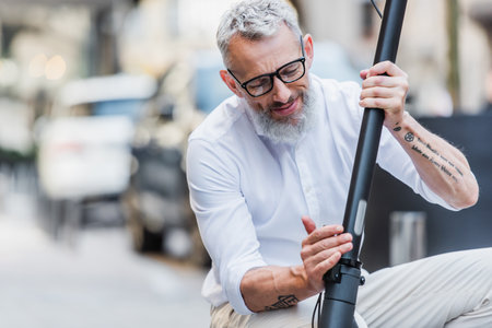 smiling and mature man in glasses adjusting electric scooter on street