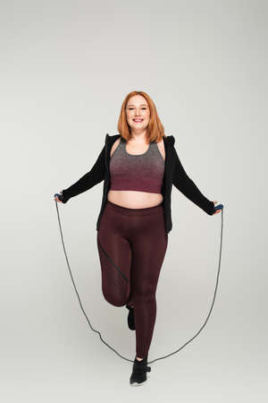 Cheerful body positive woman holding skipping rope on gray background