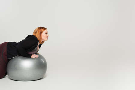 Side view of plus size woman training on fitness ball on gray background