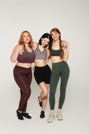 Young body positive sportswomen looking at camera and hugging on gray background