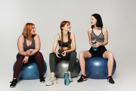 Body positive sportswoman with sports bottle looking at friends on fitness balls on gray backgroundの写真素材