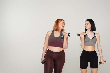 Cheerful body positive sportswomen training with dumbbells isolated on grayの写真素材